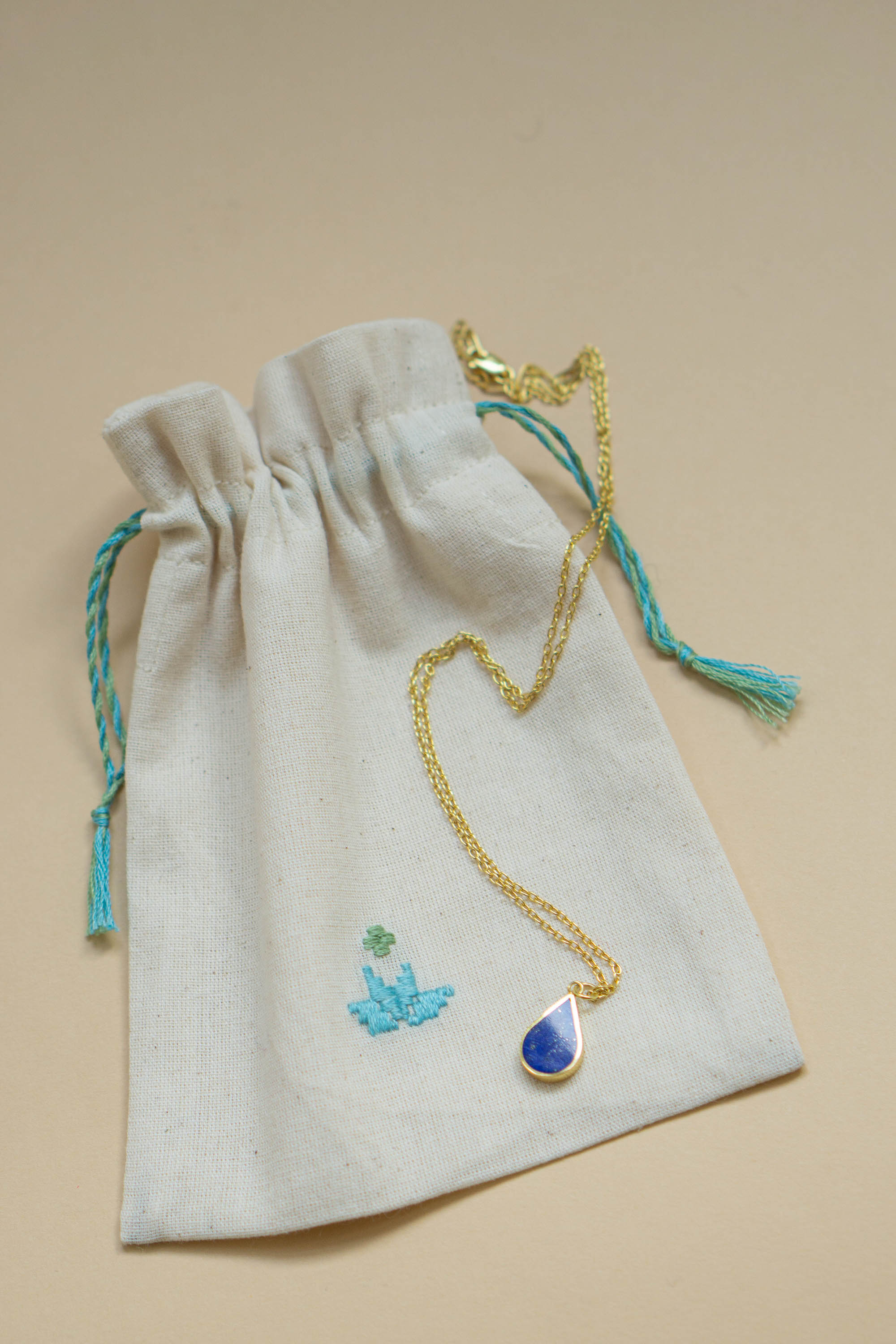Håndlavet halskæde i 24 karat forgyldt sølv med en elegant dråbeformet lapis lazuli sten, præsenteret på en broderet pose fra Afghanistan. En del af Chashma serien, inspireret af naturlige vandkilder.